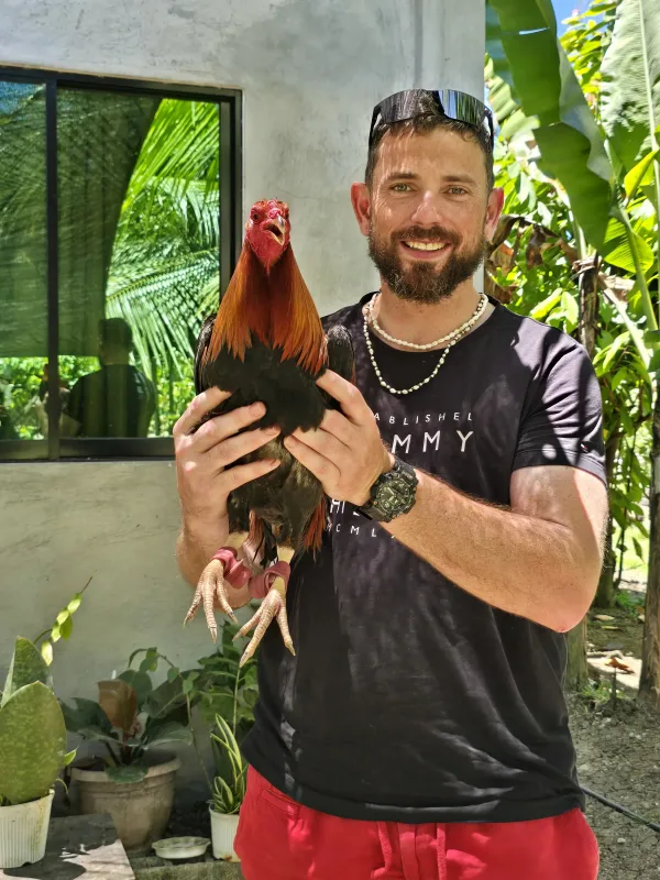 Glen O'Donnell smiling while holding a rooster among tropical plants
